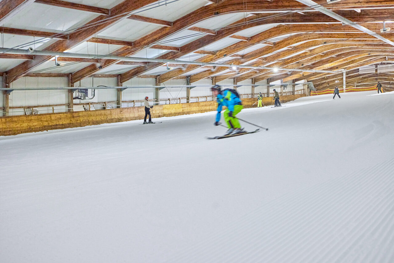 Alpincenter, Bottrop: Skifahrer in buntem Anzug sausen auf Indoor-Schneepiste mit Holzdecke und Beleuchtung.