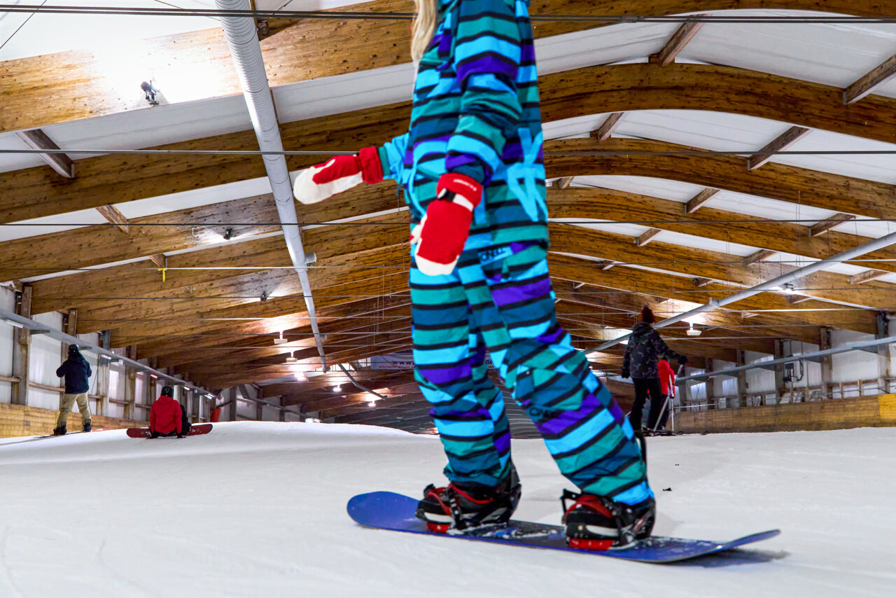 Alpincenter Bottrop: Personen fahren in einer überdachten Skihalle auf Snowboards über eine schneebedeckte Piste.