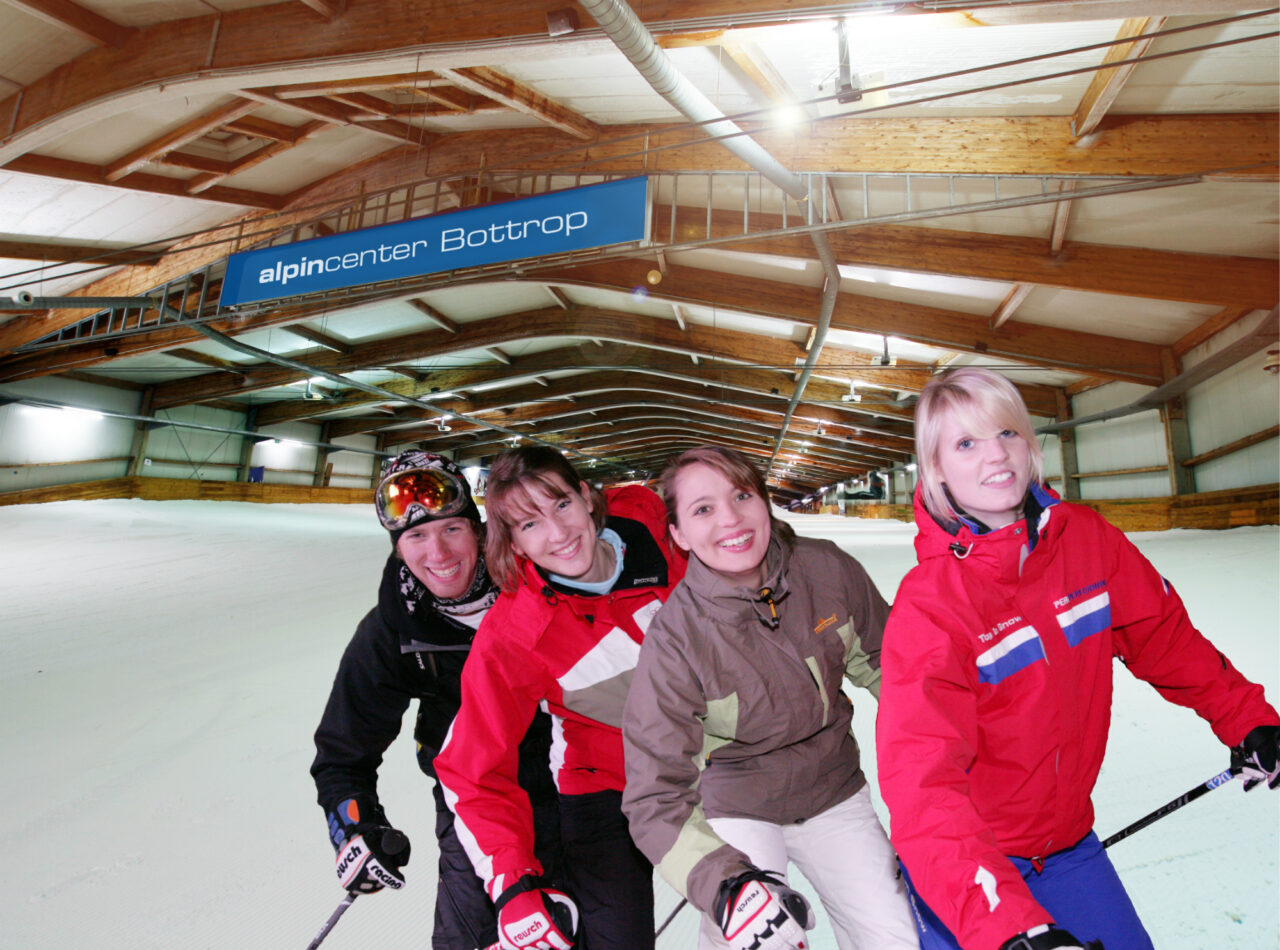 alpincenter Skihalle Piste 2111.jpg: Vier Menschen in Winterkleidung stehen lachend auf Skiern in der Skihalle des alpincenters Bottrop.