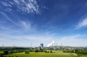 Blick vom Tetraeder, Bottrop: Industrieanlage mit rauchendem Schornstein in grüner Landschaft unter blauem Himmel.