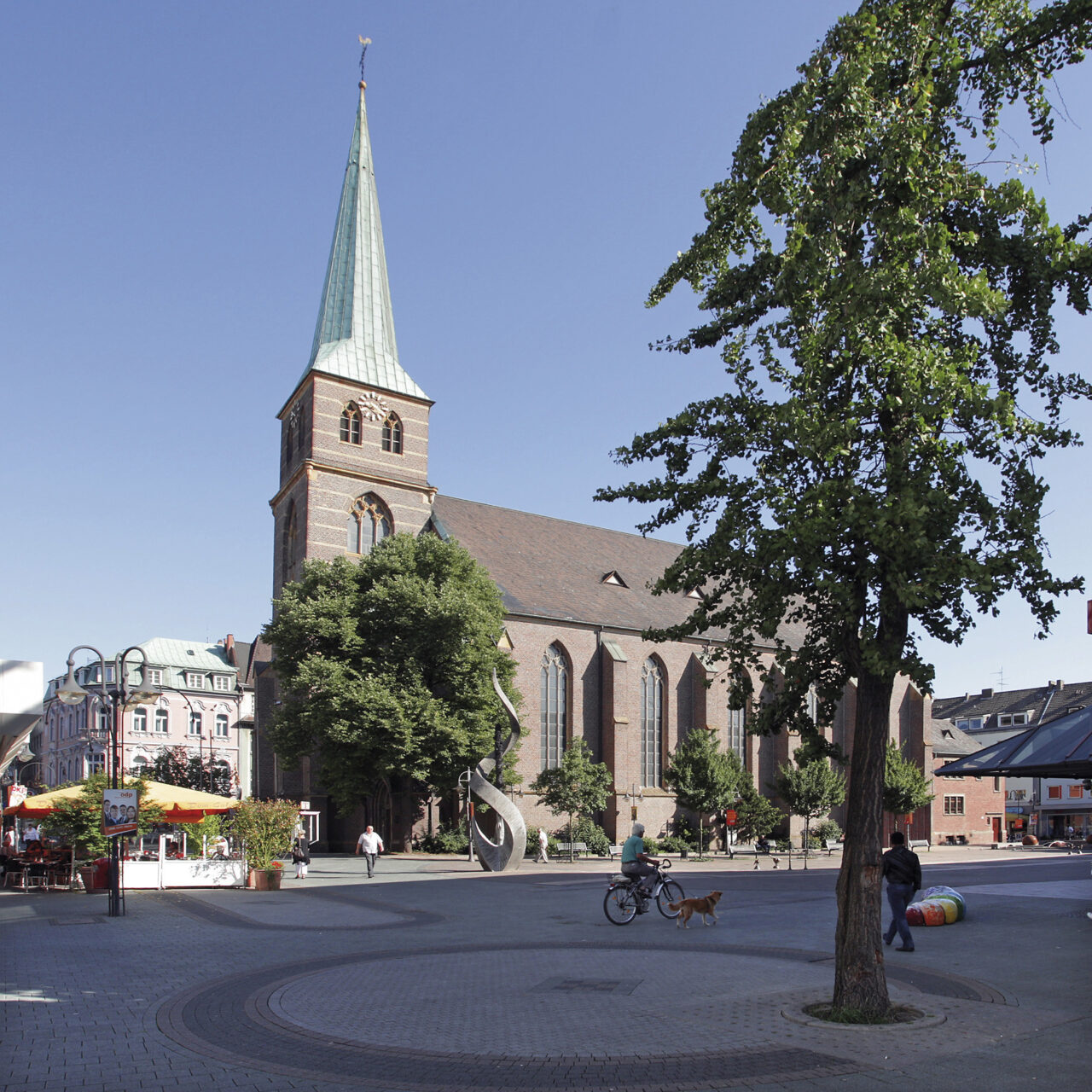 Bottrop_StCyriakus.jpg: Backsteinkirche St. Cyriakus in Bottrop mit hohem Turm und Kupferspitze auf einem sonnigen Platz.