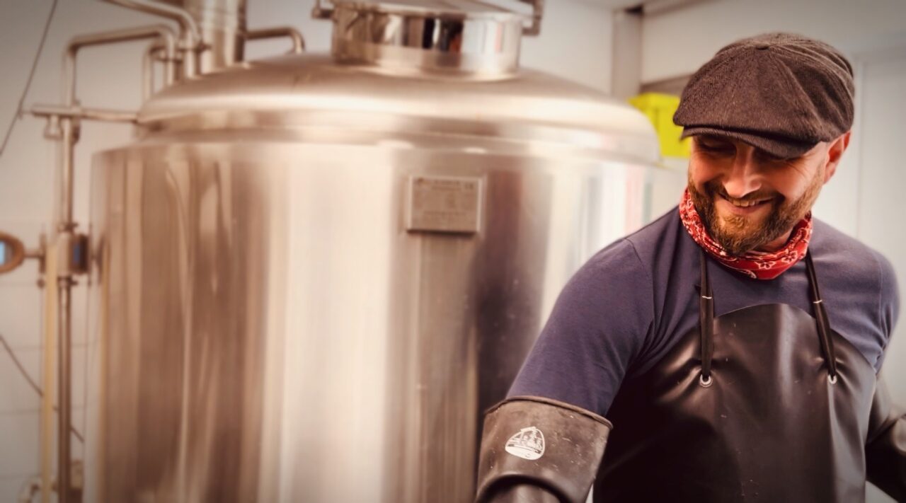 Bottroper Bier, Bottrop: Mann mit Schiebermütze lächelnd vor großem Edelstahl-Biertank in Brauerei.