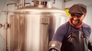 Bottroper Bier, Bottrop: Mann mit Schiebermütze lächelnd vor großem Edelstahl-Biertank in Brauerei.