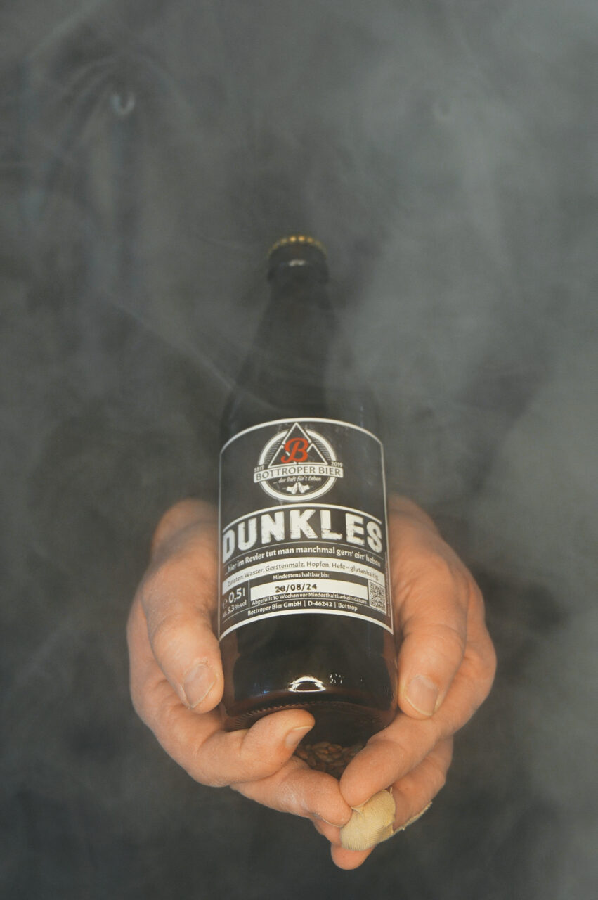 Bottroper Bier, Bottrop: Eine Person hält eine Flasche Bier "Dunkles" in rauchigem Hintergrund.