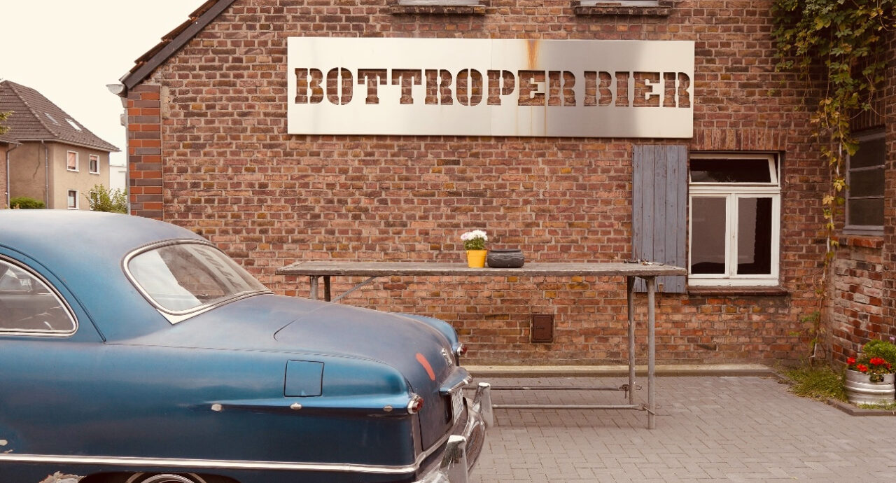 Bottroper Bier, Bottrop: Altes Gebäude mit Backsteinfassade und "Bottroper Bier"-Schild, Oldtimer daneben geparkt.
