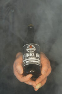 Bottroper Bier, Bottrop: Eine Person hält eine Flasche Bier "Dunkles" in rauchigem Hintergrund.
