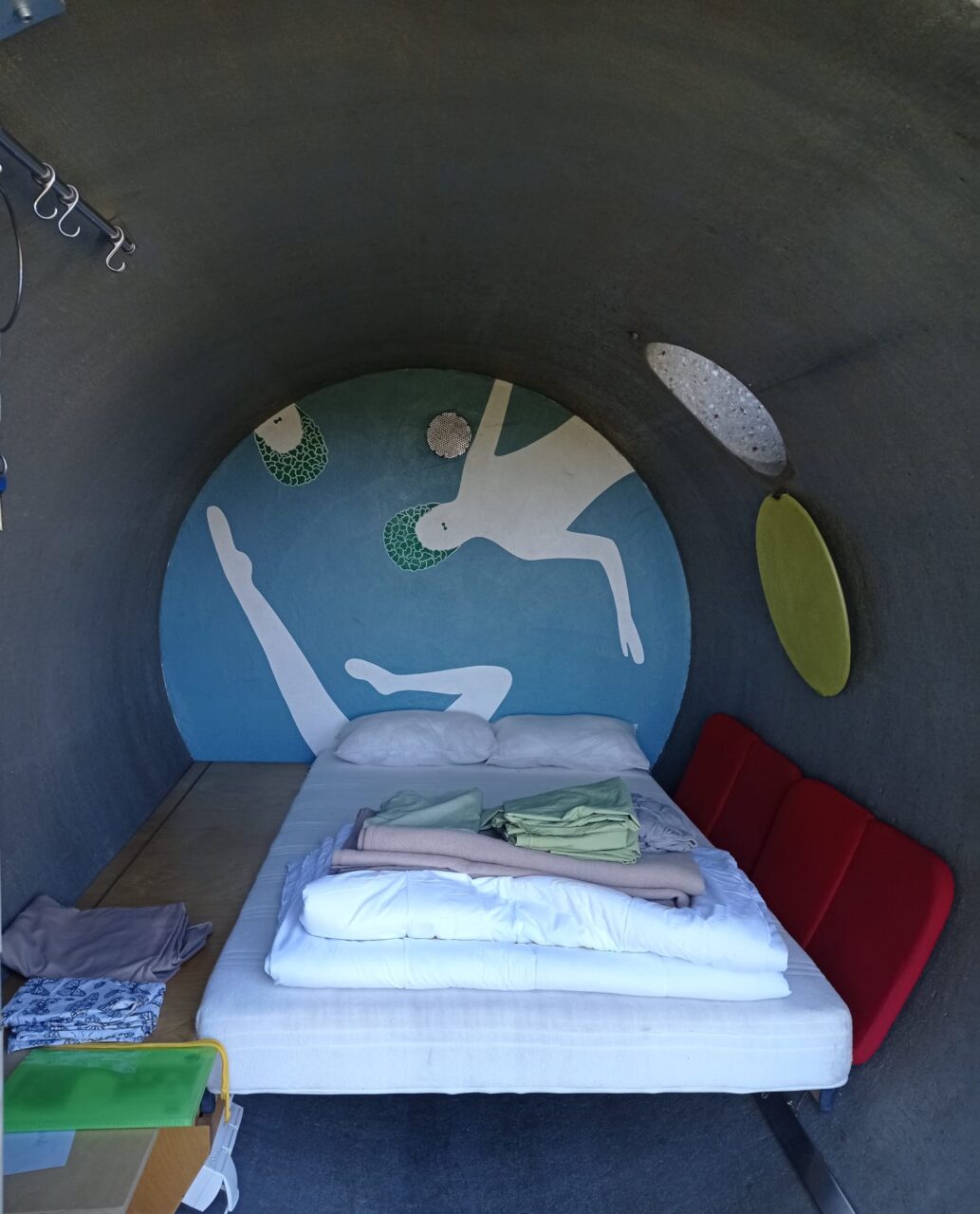 dasparkhotel BernePark, Bottrop: Innenraum eines zylindrischen Betonraums mit Bett, bunter Wandmalerei und roter Rückbank.