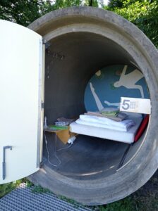 dasparkhotel BernePark, Bottrop: Rundes Betonrohr mit offener Tür, darin ein Bett mit Decken und Wandkunst im BernePark Bottrop.