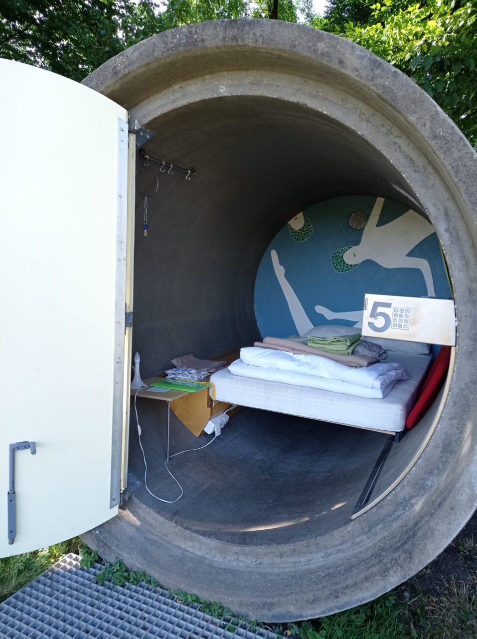 dasparkhotel BernePark, Bottrop: Rundes Betonrohr mit offener Tür, darin ein Bett mit Decken und Wandkunst im BernePark Bottrop.