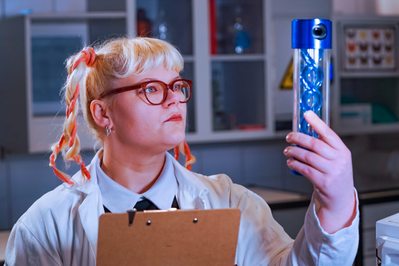 ELORIA Erlebnisfabrik, Bottrop: Person im weißen Laborkittel betrachtet in einem Labor aufmerksam ein blaues Objekt in einem zylindrischen Behälter.