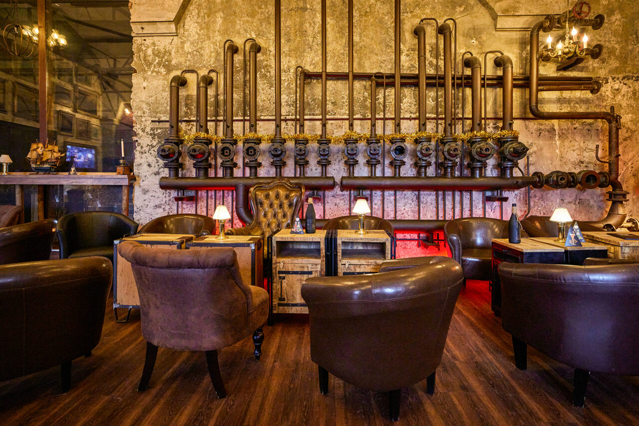 ELORIA Erlebnisfabrik Bottrop: Eine industriell gestaltete Bar mit Ledersesseln und alten Rohrleitungen an der Wand.