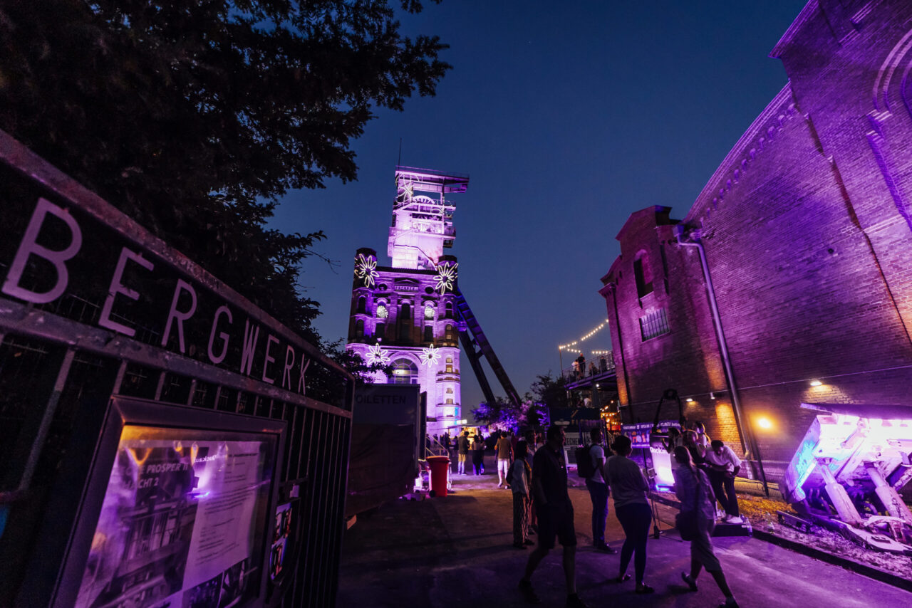 ExtraSchicht, Malakoffturm Zeche Prosper II, Bottrop: Abendstimmung vor historischer Zechenanlage, illuminiert in violettem Licht, bei einer Veranstaltung.