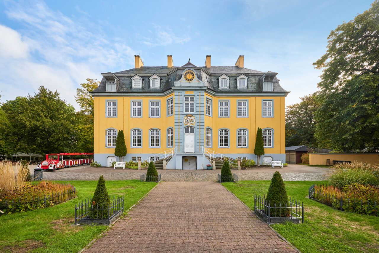 Freizeitpark Schloss Beck: Gelbes Barockschloss mit grauem Dach und Gartenweg, links eine rote Parkeisenbahn vor Bäumen.