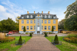 Freizeitpark Schloss Beck: Gelbes Barockschloss mit grauem Dach und Gartenweg, links eine rote Parkeisenbahn vor Bäumen.