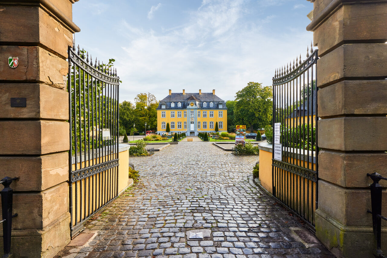 Freizeitpark Schloss Beck, Bottrop: Blick durch geöffnete Torflügel auf das gelbe Schloss Beck mit gepflastertem Hof und Gartenanlage.
