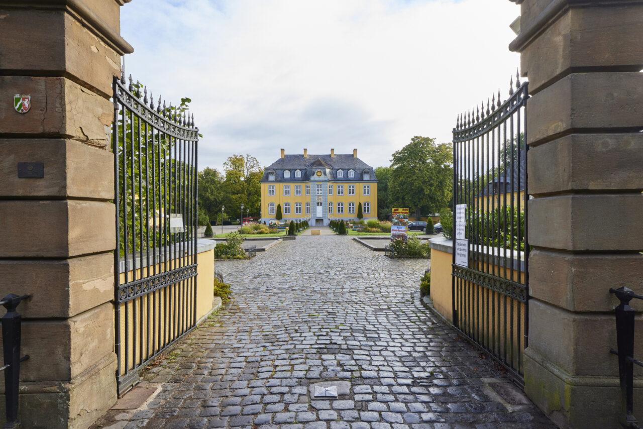 Freizeitpark Schloss Beck, Bottrop: Ein barockes, gelbes Schloss hinter einem offenen Tor mit gepflastertem Hof im Vordergrund.