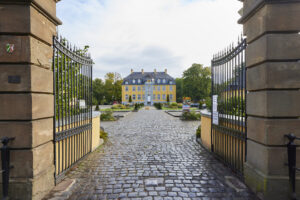 Freizeitpark Schloss Beck, Bottrop: Ein barockes, gelbes Schloss hinter einem offenen Tor mit gepflastertem Hof im Vordergrund.