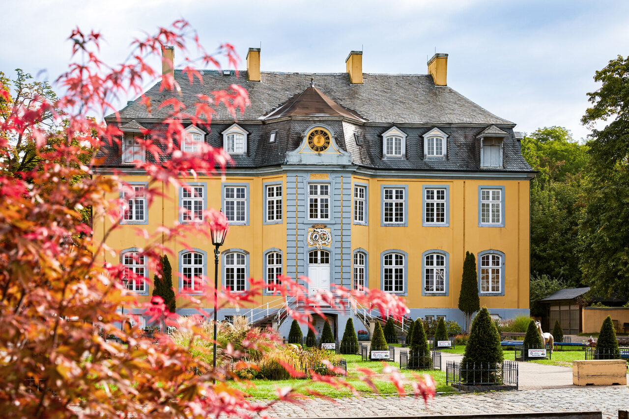 Freizeitpark Schloss Beck Bottrop: Historisches gelbes Gebäude mit grauen Fensterläden, flankiert von grünem Laub im Vordergrund.