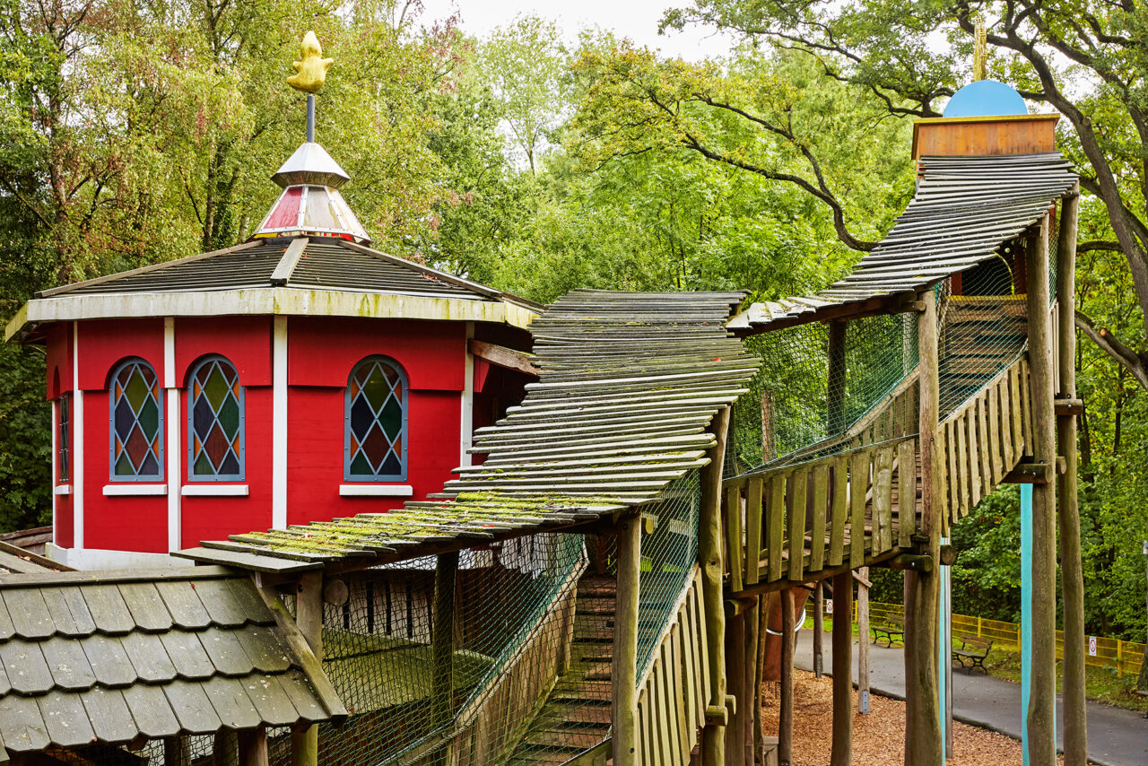 Freizeitpark Schloss Beck Bottrop: Ein farbenfrohes Spielhaus mit gewellter Holzbrücke und umliegenden Bäumen im Park.