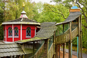 Freizeitpark Schloss Beck Bottrop: Ein farbenfrohes Spielhaus mit gewellter Holzbrücke und umliegenden Bäumen im Park.