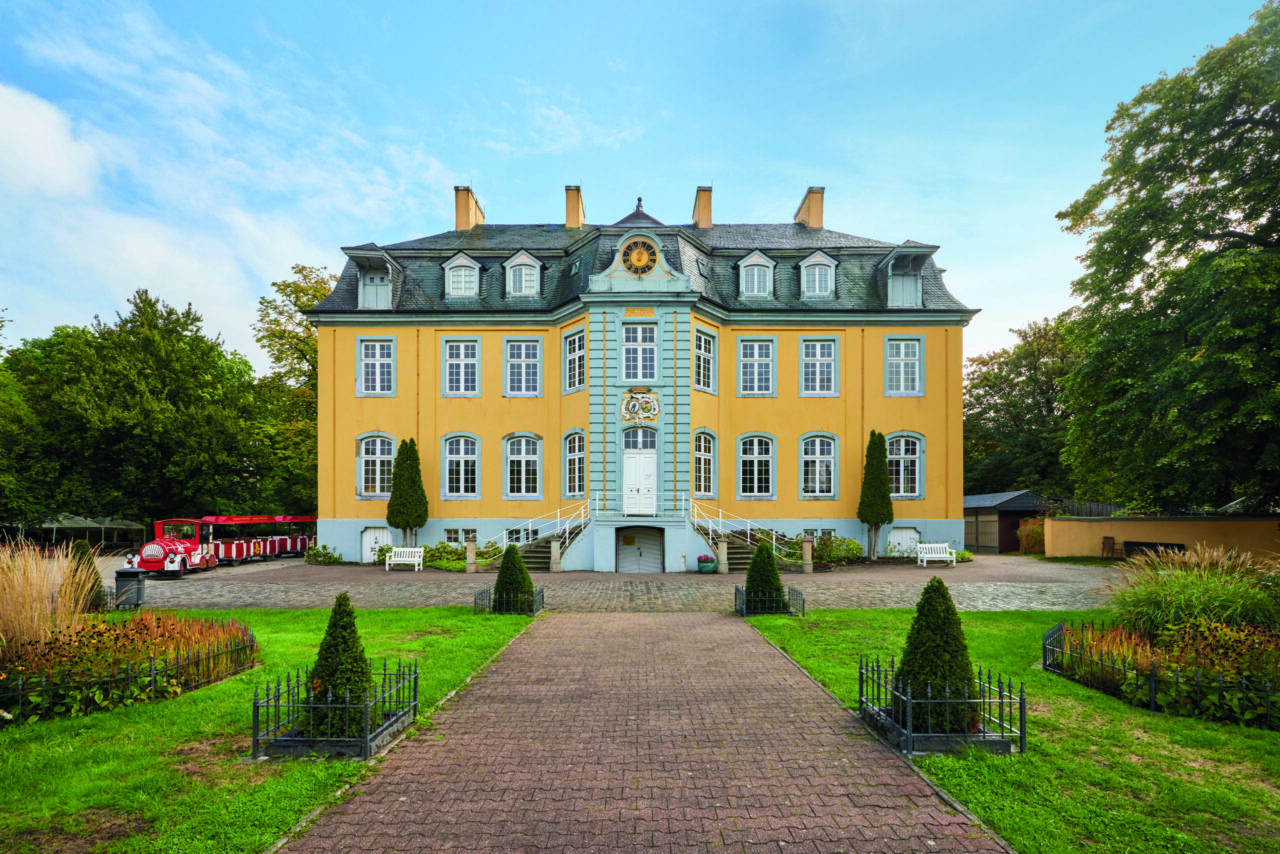 Freizeitpark Schloss Beck, Bottrop: Gelbes Barockschloss mit symmetrischer Fassade, Gartenweg und roter Parkbahn im Vordergrund