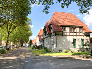 Gartenstadt Welheim, Bottrop: Baumgesäumte Straße mit historischen Wohnhäusern der Gartenstadt Welheim unter blauem Himmel.