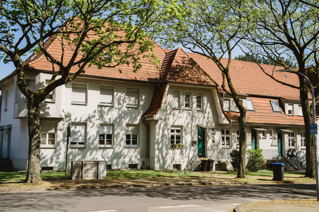 Gartenstadt Welheim, Bottrop: Haus im Grünen mit roten Ziegeldächern und umliegenden Bäumen, sonnendurchflutete Straße im Vordergrund.