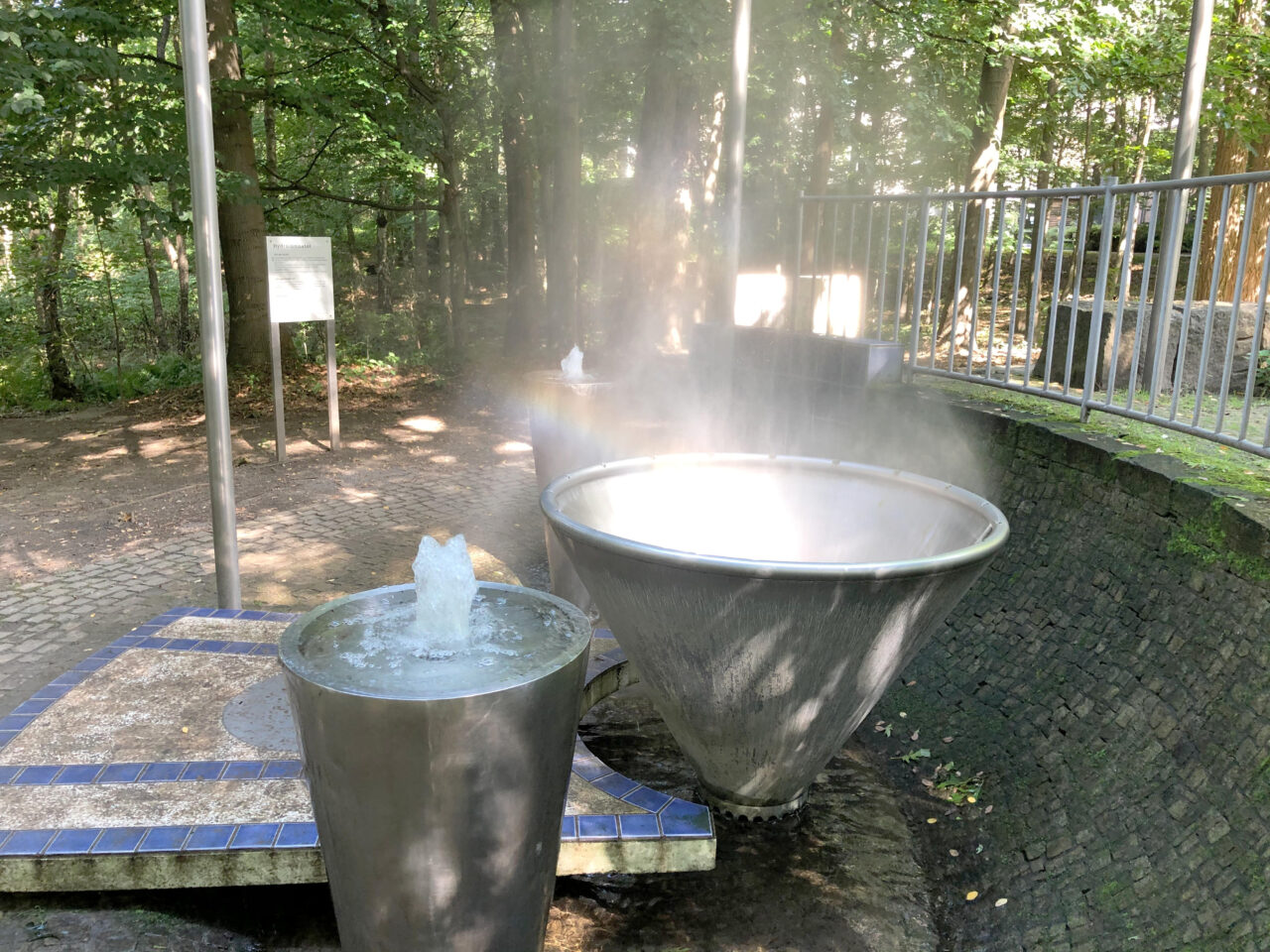 Gesundheitspark Quellenbusch, Bottrop, Brunnen: Metallene Brunnenanlage mit aufsteigendem Dampf, umgeben von Bäumen im Gesundheitspark Bottrop.