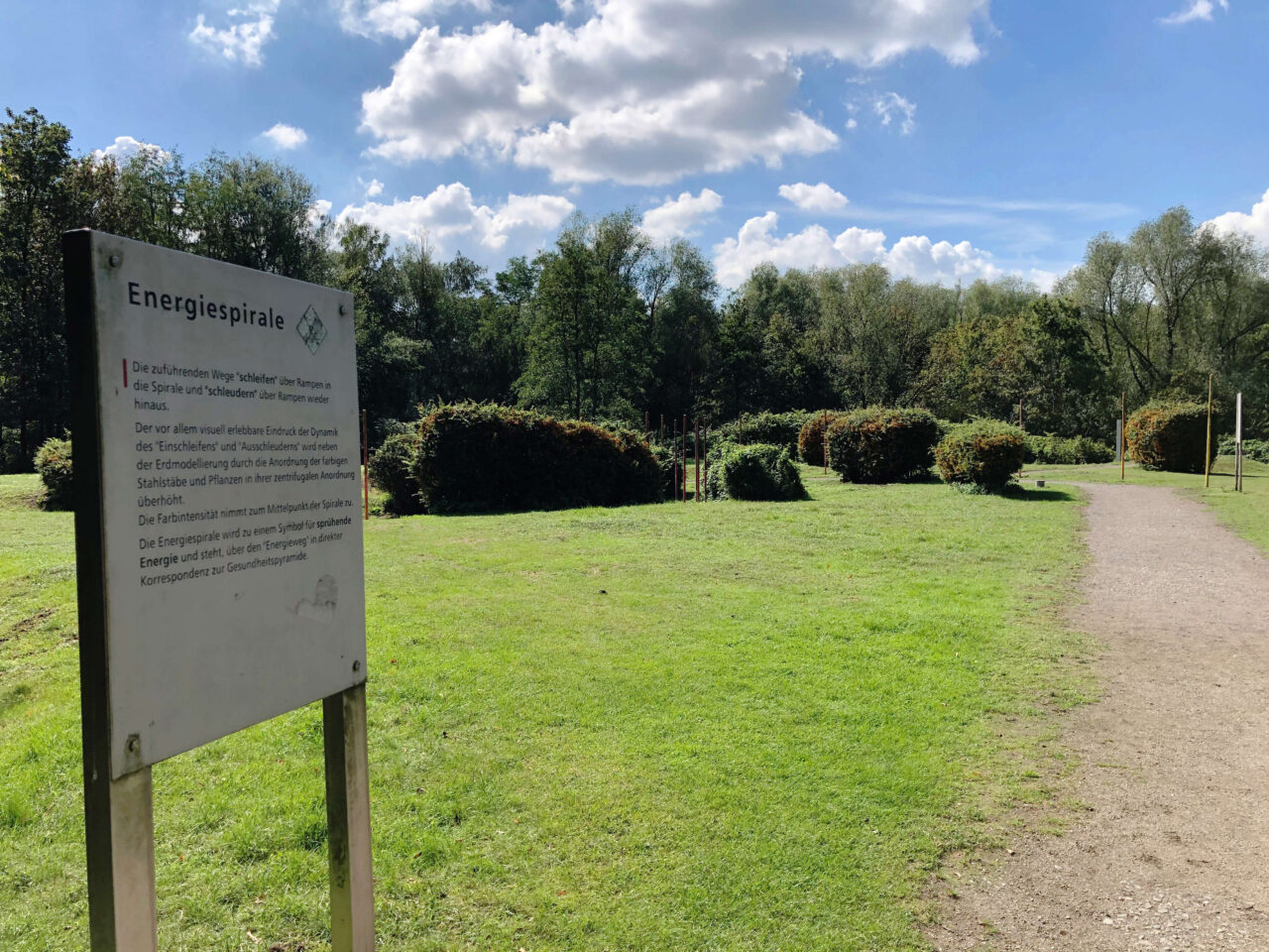 Gesundheitspark Quellenbusch, Bottrop, Weg und Schild: Hinweisschild zur Energiespirale im Gesundheitspark Quellenbusch mit Weg und grüner Wiesenfläche