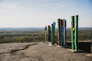 Halde Haniel, Bottrop: Bunte Holzstelen stehen auf einem Hügel und bieten einen weiten Blick über eine bewaldete Landschaft.