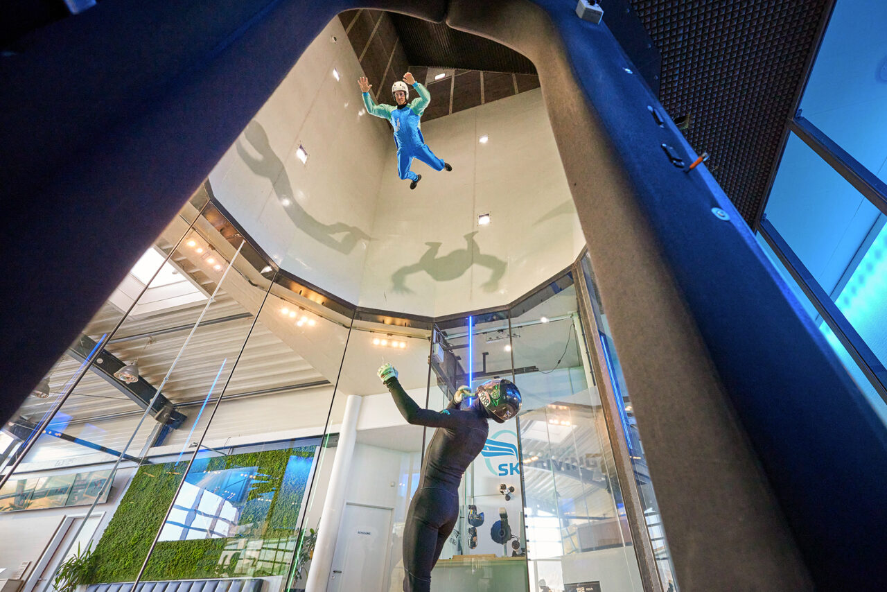 Indoor Skydiving Bottrop: Indoor-Skydiving: Zwei Personen schweben in einem vertikalen Windkanal, umgeben von Glaswänden.