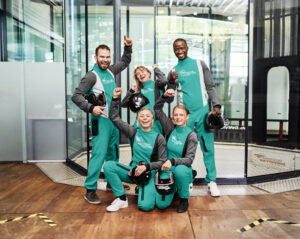 Indoor Skydiving, Bottrop, Freunde: Gruppe lachender Menschen in grünen Anzügen posiert in der Halle des Indoor Skydiving Bottrop.