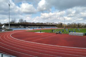 Jahnstadion Bottrop: Fußballstadion mit Laufbahn, überdachter Tribüne und Trainierenden auf dem Rasen unter Wolkenhimmel.