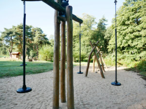 Kirchheller Heide, Bottrop, Waldspielplatz: Holzspielgeräte mit Schaukeln im Sandbereich, umgeben von grünen Wiesen und Bäumen der Kirchheller Heide.