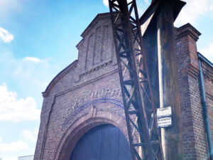 Lokschuppen, Bottrop, Außenansicht vorne: Backsteinbau mit großem Rundbogenportal und Metallträger vor der Fassade unter blauem Himmel