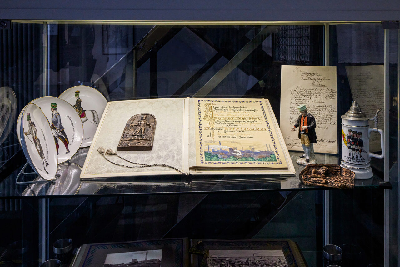 Malakoffturm, Bottrop: Glasvitrine mit handschriftlichen Dokumenten, Bierkrug, Zinnfigur und dekorativen Tellern.
