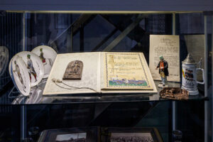 Malakoffturm, Bottrop: Glasvitrine mit handschriftlichen Dokumenten, Bierkrug, Zinnfigur und dekorativen Tellern.