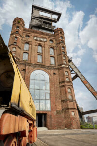 Malakoffturm, Bottrop: ```html
Alte Zeche Prosper, roter Backsteinbau mit hohen Fenstern und Förderturm, im Vordergrund ein Zug.
```