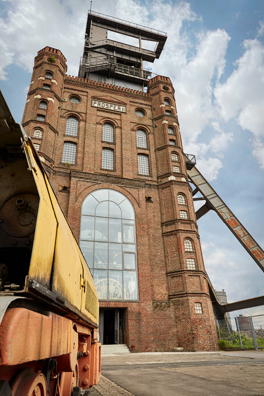 Malakoffturm, Bottrop: ```html
Alte Zeche Prosper, roter Backsteinbau mit hohen Fenstern und Förderturm, im Vordergrund ein Zug.
```