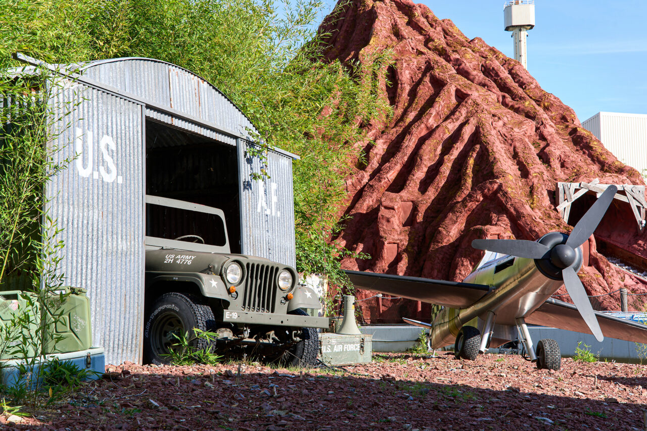 Movie Park Germany Bottrop: Ein Militärjeep steht halb in einem Blechschuppen; daneben ein Flugzeug mit Vulkankulisse.