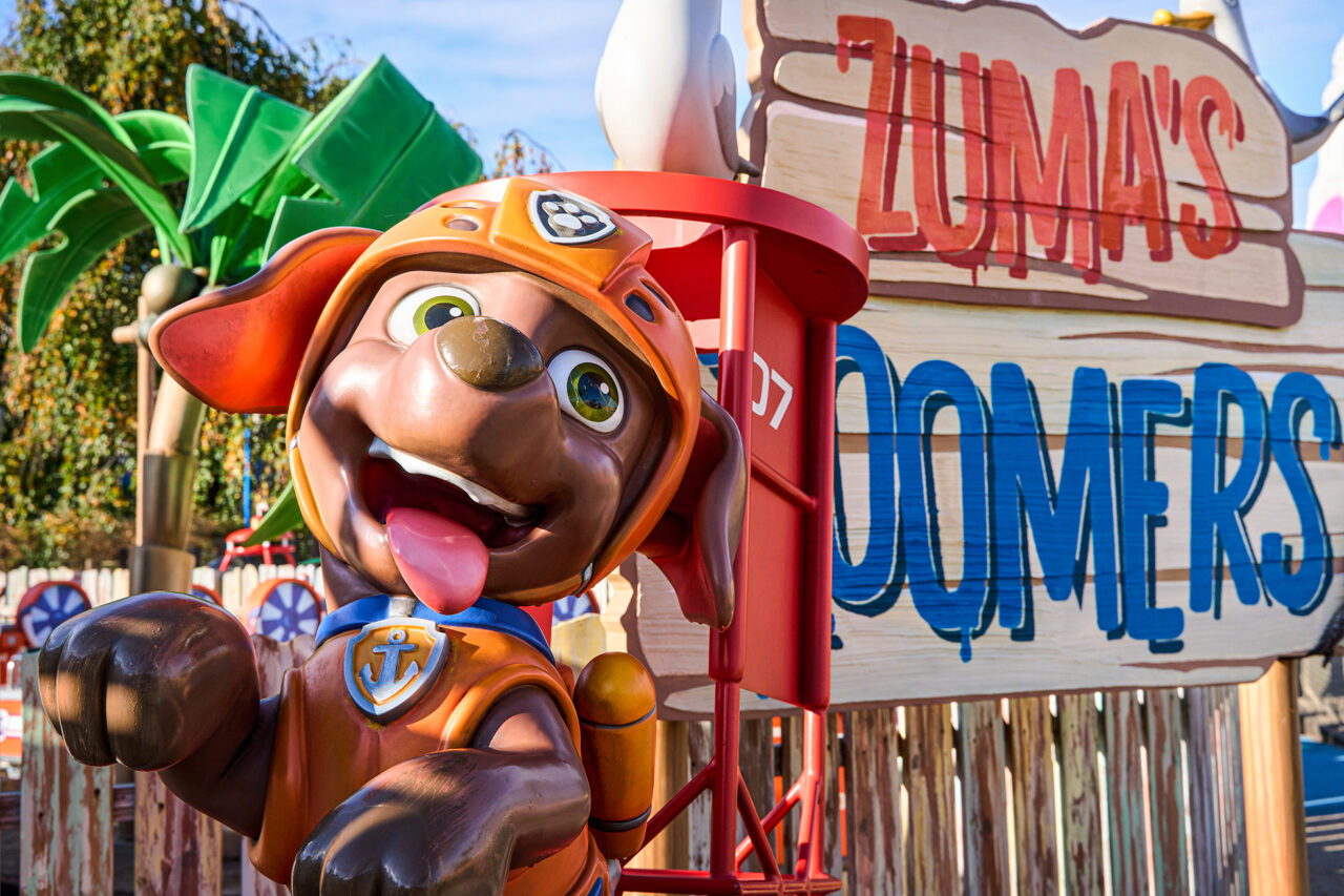Movie Park Germany Bottrop: Fröhliche Figur von Zuma aus Paw Patrol vor dem bunten Schild "Zuma's Zoomers" im Freizeitpark.