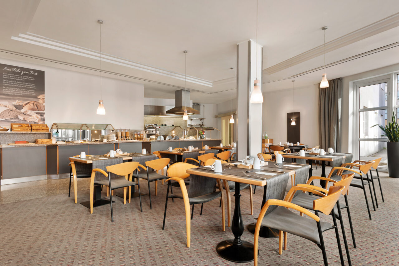 Ramada Bottrop - Breakfast - 1354613.jpg: Helles Frühstücksrestaurant mit Holztischen, modernem Buffetbereich und großen Fenstern im Ramada Bottrop