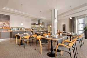 Ramada Bottrop - Breakfast - 1354613.jpg: Helles Frühstücksrestaurant mit Holztischen, modernem Buffetbereich und großen Fenstern im Ramada Bottrop