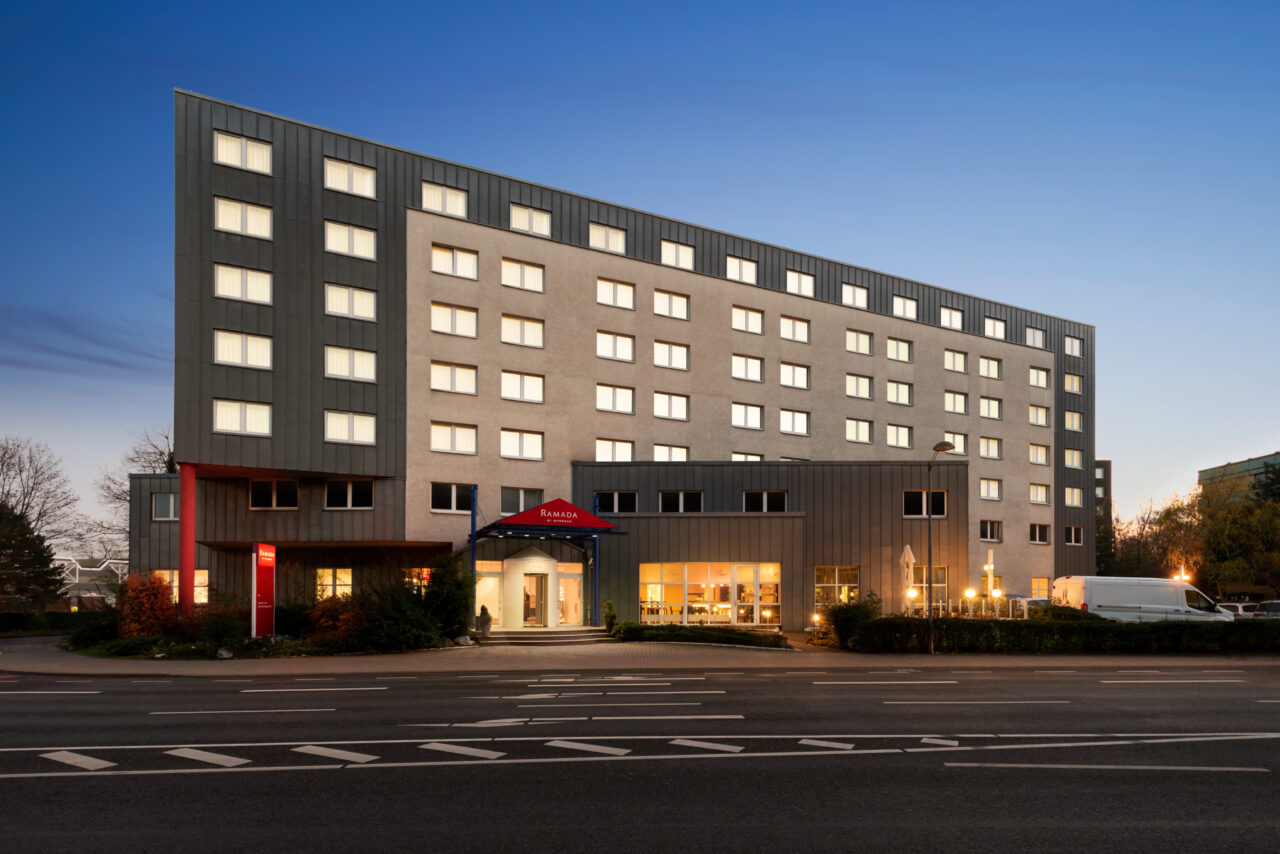Ramada Bottrop - Exterior - 1354621.jpg: Modernes sechsstöckiges Hotelgebäude mit beleuchteten Fenstern und Eingang bei Abenddämmerung