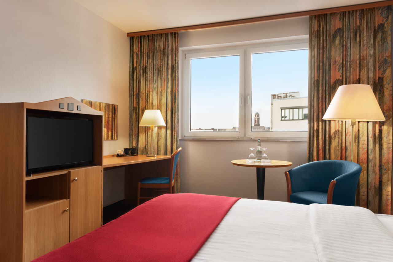Ramada Bottrop - Standard Double Bed - 1354602.jpg: Hotelzimmer mit Doppelbett, roter Decke, Schreibtisch, Fernseher und Fenster mit Stadtblick.