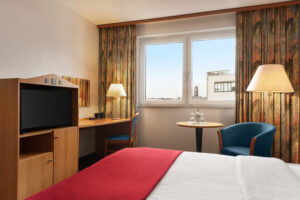 Ramada Bottrop - Standard Double Bed - 1354602.jpg: Hotelzimmer mit Doppelbett, roter Decke, Schreibtisch, Fernseher und Fenster mit Stadtblick.