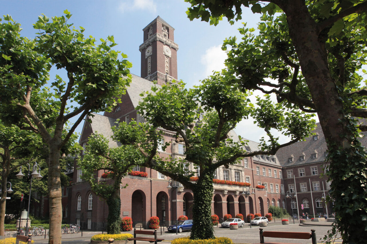 Rathaus, Bottrop, Blick zum Turm: Backsteinernes Rathaus Bottrop mit Uhrturm, begrüntem Vorplatz und blühenden Balkonkästen im Sommer.