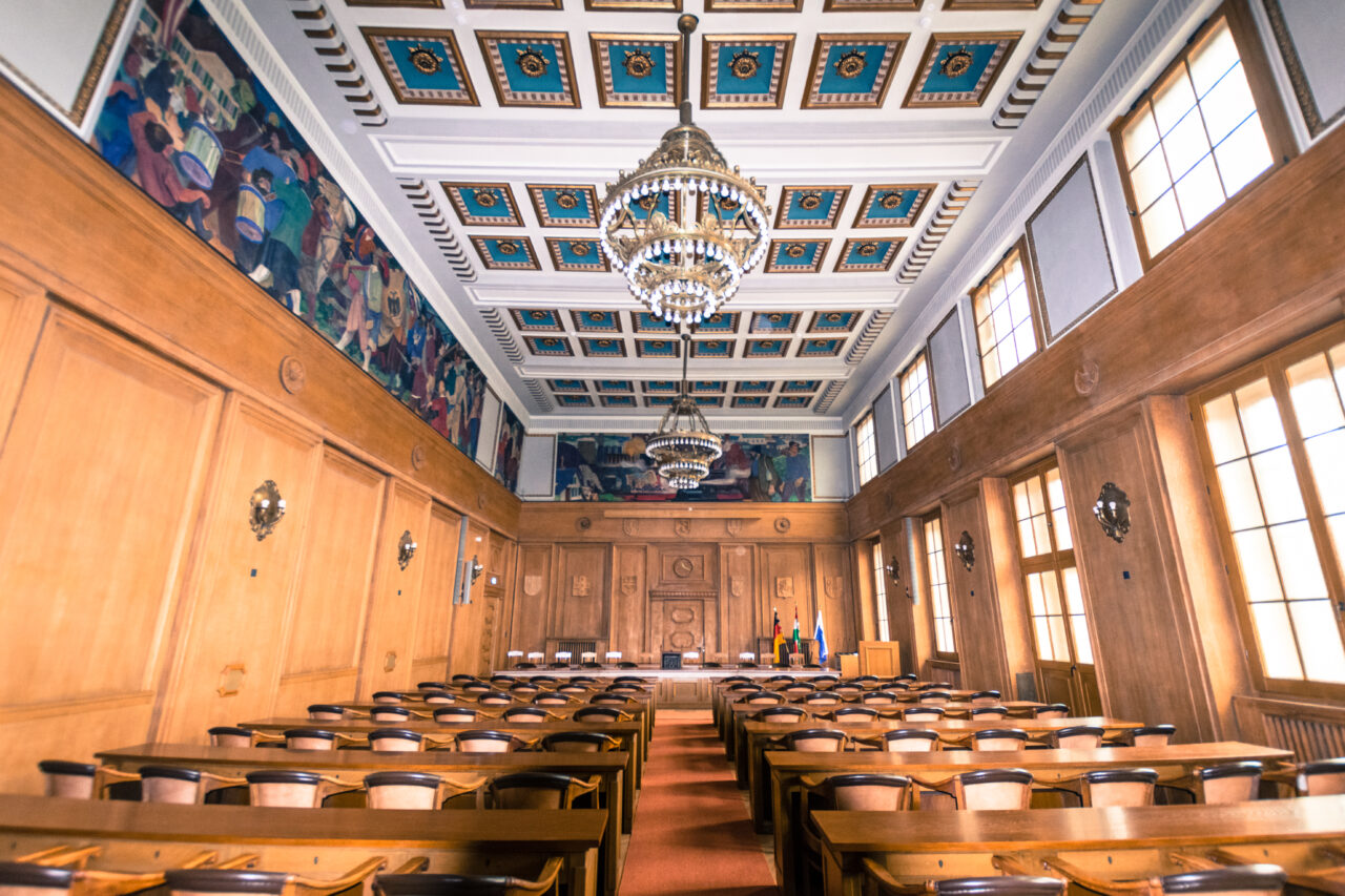 Rathaus, Bottrop, Sitzungssaal: Holzgetäfelter Sitzungssaal im Rathaus Bottrop mit Kronleuchtern, Wandgemälden und hohen Fenstern.