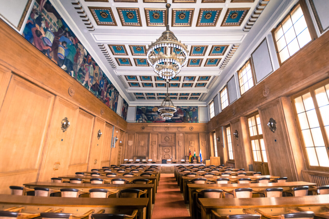 Rathaus, Bottrop, Sitzungssaal: Holzgetäfelter Sitzungssaal im Rathaus Bottrop mit Kronleuchtern, Wandgemälden und hohen Fenstern.