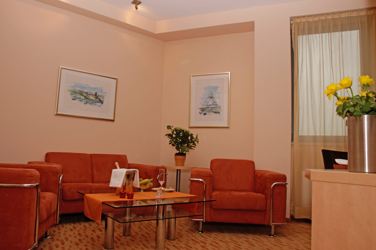 Rhein-Ruhr-Hotel, Bottrop: Sitzbereich mit orangefarbenen Polstermöbeln, Glastisch, Pflanzen und Wandbildern in warmem Licht